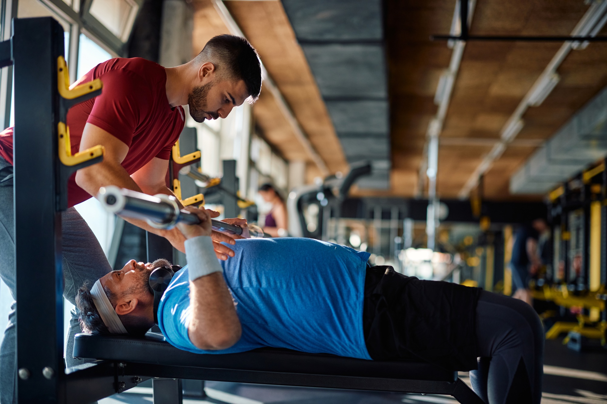 Mature man exercising with barbel with help of coach in a gym.