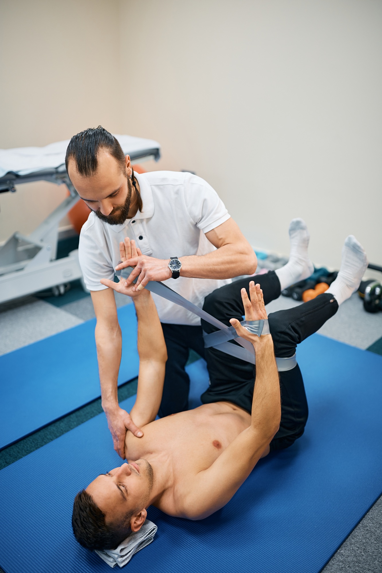 Athletic man exercising resistance band during kinesiotherapy treatment with physiotherapist.